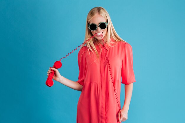 Portrait d'une fille blonde élégante à lunettes de soleil avec fil téléphonique dans la bouche isolée sur un mur bleu