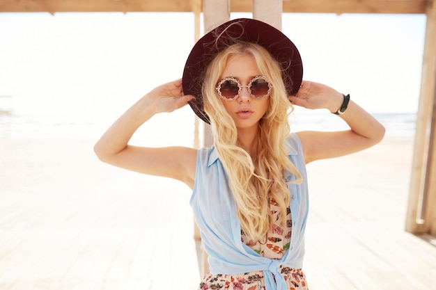 Portrait d'une fille blonde avec des lunettes de soleil jolie robe cheveux ondulés et chapeau bordeaux le fond de la lumière du soleil