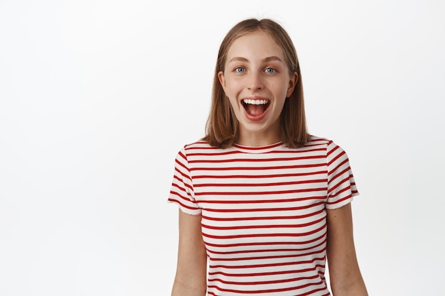 Portrait d'une fille blonde à la recherche d'étonnement et de surprise, fascinée par une grosse affaire promotionnelle, la bouche ouverte avec admiration et regardant la caméra, souriant largement, debout sur fond blanc