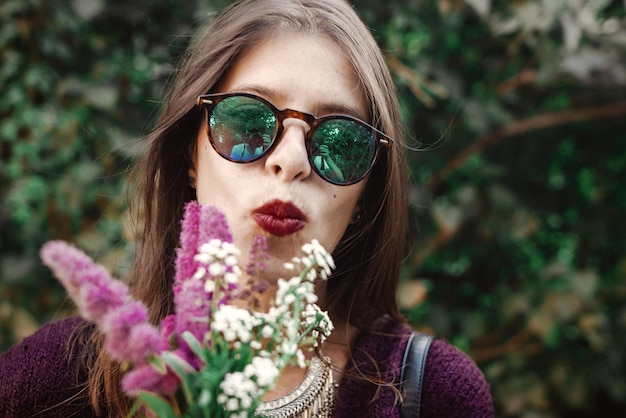 Portrait d'une fille boho heureuse en lunettes de soleil souriante avec bouquet de fleurs sauvages dans un jardin ensoleillé Fille insouciante hipster élégante s'amusant au buisson vert dans la rue Vacances d'été et voyage