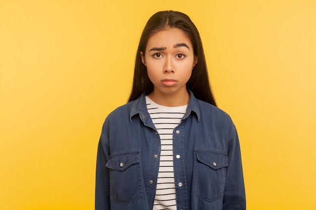 Portrait d'une fille bouleversée en chemise en jean regardant la caméra avec une grimace sombre et mécontente, se sentant agacée et insatisfaite de la défaite, des émotions négatives. tourné en studio isolé sur fond jaune