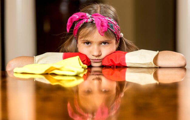 Portrait de fille bouleversée dans des gants en caoutchouc rouge polissant la table en bois