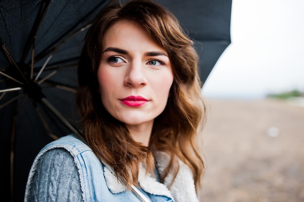 Portrait de fille brune bouclée en veste jeans avec parapluie noir sur le terrain