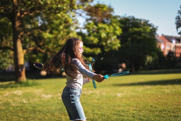 Le portrait d'une fille caucasienne souffle des bulles de savon encerclant dans le parc sur un pré de terrain de jeu