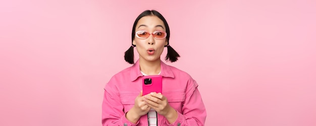 Portrait d'une fille coréenne dans des lunettes de soleil élégantes tenant un téléphone portable à l'aide d'une application pour smartphone debout au-dessus