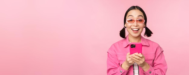 Portrait d'une fille coréenne dans des lunettes de soleil élégantes tenant un téléphone portable à l'aide d'une application pour smartphone debout sur fond rose