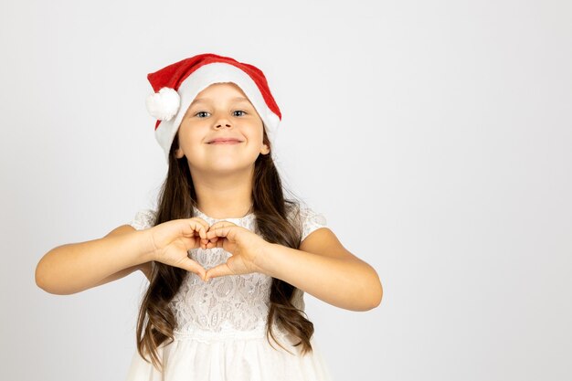 Portrait d'une fille douce et heureuse en robe blanche portant un chapeau de père Noël rouge montre un signe de coeur de doigt...