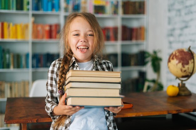 Portrait d'une fille de l'école élémentaire joyeuse tenant une pile de livres dans la bibliothèque à l'école à la recherche