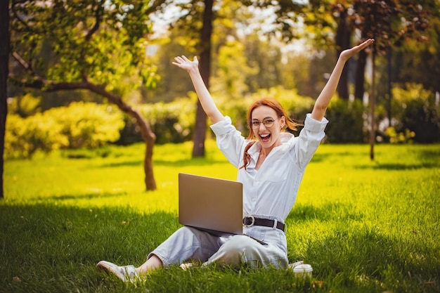 Portrait d'une fille émotionnelle joyeuse assise dans le parc tout en utilisant un ordinateur portable s'amusant à se réjouir à l'extérieur