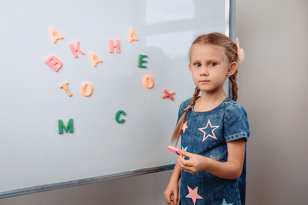 Portrait d'une fille enfant assez perplexe debout en face d'un grand tableau blanc sur lequel les lettres de l'alphabet sont dispersées de manière chaotique. Concept de connaissances photo avec bruit