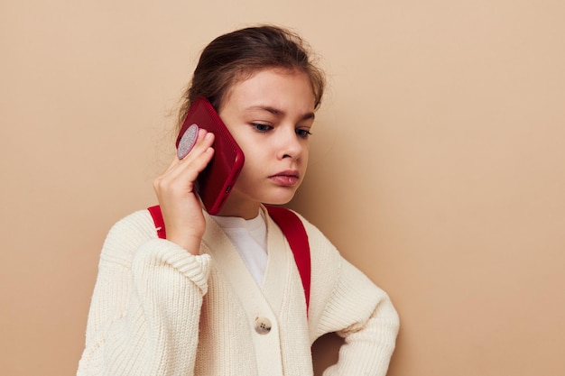 Portrait d'une fille enfant souriante heureuse avec un téléphone posant un fond isolé de sac à dos rouge