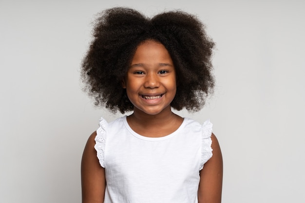 Portrait d'une fille frisée d'âge préscolaire heureuse en T-shirt regardant joyeusement la caméra et souriante, fière de son propre succès. Studio intérieur tourné isolé sur fond blanc