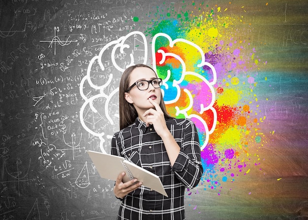 Photo portrait d'une fille geek en lunettes debout près d'un tableau noir avec un cahier et un grand cerveau coloré avec des formules autour de lui.