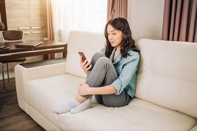 Portrait d'une fille heureuse assise dans un canapé avec un téléphone portable, connectée au wifi ayant un appel vidéo, lisant des commentaires agréables sous sa photo dans les réseaux sociaux, vêtue de vêtements de maison décontractés. Intérieur confortable.