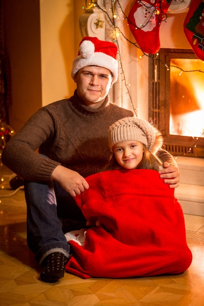 Portrait De Fille Heureuse Dans Le Sac De Santa Assis Avec Le Père à Côté De La Cheminée