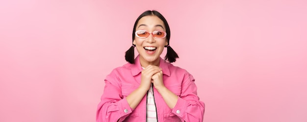 Portrait d'une fille japonaise excitée dans des lunettes de soleil célébrant l'atteinte de l'objectif haletant étonné et souriant debout sur fond rose