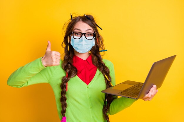 Portrait d'une fille joyeuse et funky tenant dans les mains un ordinateur portable montrant le pouce vers le haut de la publicité porter un masque isolé sur fond de couleur jaune