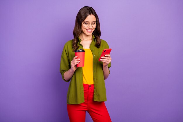 Portrait d'une fille joyeuse et positive utilisant un smartphone en tapant sur le chat en train de se rafraîchir tenir à emporter tasse à latte porter un style élégant pantalon rouge à la mode pantalon isolé sur fond de couleur violet