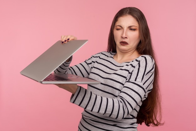 Portrait d'une fille mécontente en sweat-shirt rayé fermant un ordinateur portable, regardant avec suspicion et méfiance à l'écran, effrayée par un logiciel inconnu, un échec, un bogue de programme. studio tourné en intérieur, fond rose