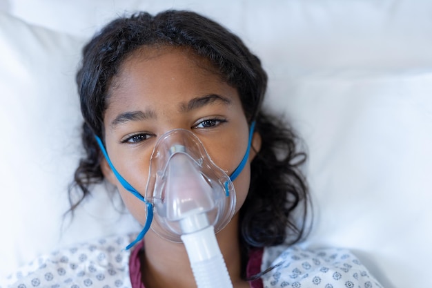 Portrait d'une fille métisse malade allongée dans un lit d'hôpital portant un ventilateur à masque à oxygène