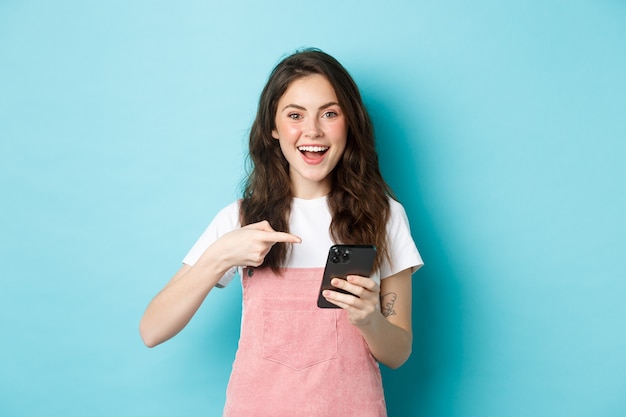 Portrait d'une fille mignonne et joyeuse pointant le doigt sur son téléphone, à l'aide d'une application d'achat pour smartphone, debout sur fond bleu