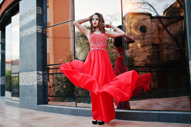 Portrait d'une fille à la mode à la robe de soirée rouge posée fenêtre miroir de fond du bâtiment moderne. Robe soufflant dans l'air