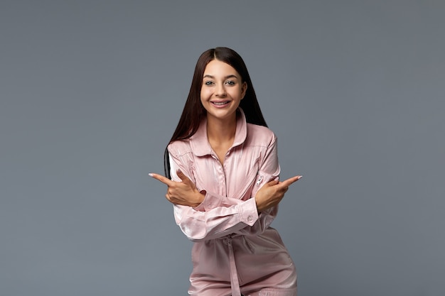 Portrait d'une fille de mode souriante en salopette rose pointant son doigt vers l'espace de copie vide. Copiez l'espace, fond gris. Concept de mode.