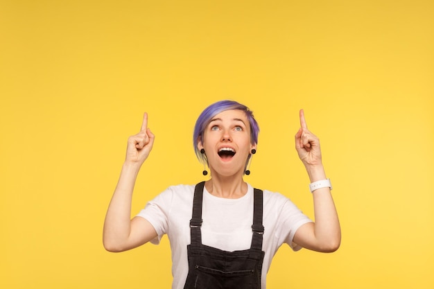 Portrait d'une fille à la mode surprise avec des cheveux courts violets en salopette en jean pointant vers l'espace libre pour la publicité, l'air choqué. isolé sur fond jaune, prise de vue en studio, espace de copie