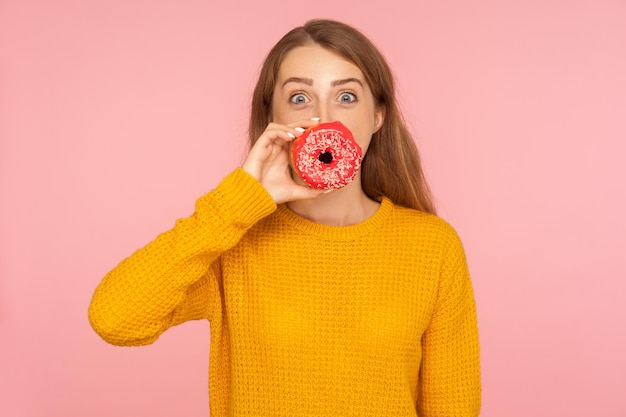 Portrait d'une fille positive aux cheveux roux en pull couvrant la bouche avec un beignet et regardant étonnée devant la caméra, mettant un beignet rose sur les lèvres, s'amusant avec des aliments sucrés. tourné en studio isolé sur fond rose