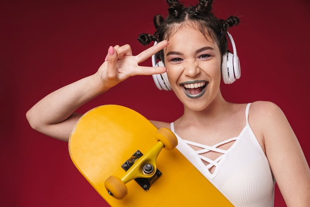 Portrait D'une Fille Punk Optimiste Avec Une Coiffure Bizarre Et Un Rouge à Lèvres Foncé Tenant Une Planche à Roulettes Tout En écoutant De La Musique Avec Des écouteurs Isolés Sur Un Mur Rouge