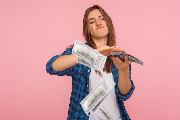 Portrait d'une fille riche et gaspilleuse en chemise à carreaux éparpillant des dollars avec une grimace arrogante, se vantant d'une vie riche, concept de dépenses d'argent imprudentes. studio d'intérieur tourné isolé sur fond rose