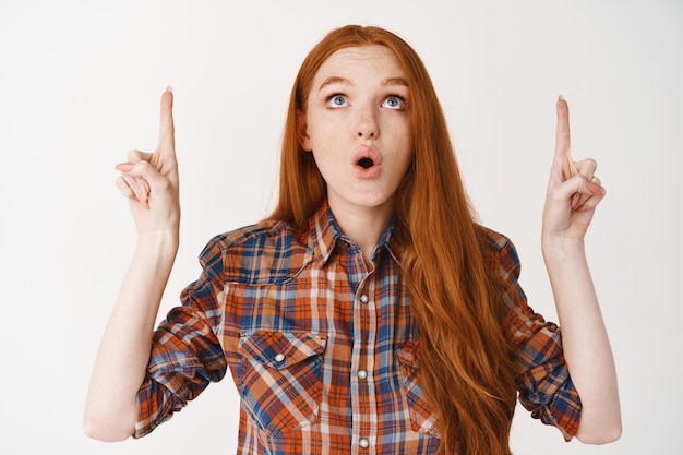 Portrait d'une fille rousse étonnée pointant les doigts vers le haut, regardant le logo avec intérêt, debout sur un mur blanc