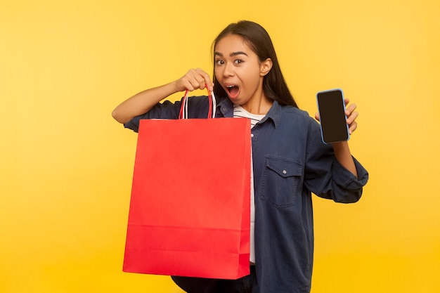 Portrait d'une fille shopper ravie et étonnée en chemise en jean tenant des sacs et un téléphone portable avec une maquette d'affichage vide, surprise par des achats fous dans un magasin de mode, une vente d'occasions. tourné en studio intérieur isolé