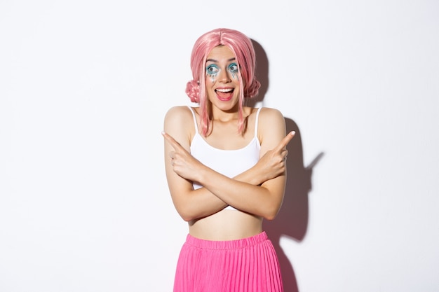 Portrait d'une fille souriante excitée avec une perruque rose et un maquillage de fête, pointant les doigts sur le côté mais regardant à gauche avec une expression heureuse, debout sur fond blanc.