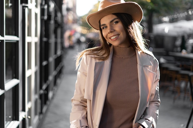 Portrait d'une fille souriante à la mode dans la rue d'une ville européenne avec des cheveux lâches et de belles dents blanches dans un chapeau rétro et un manteau beige. Modèle dans la rue.