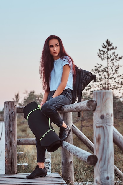 Portrait d'une fille touristique en chemise blanche et jeans s'appuyant sur une clôture en bois dans une belle prairie d'automne au coucher du soleil.