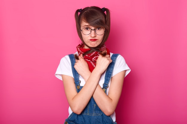 Portrait de fille triste porte un t-shirt blanc, une salopette en jean, avec un bandana sur le cou. Écolière croise les mains sur la poitrine, tient des nattes, a l'air bouleversé, ne veut pas retourner à l'école après les vacances.