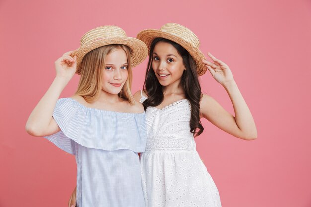 Portrait de filles d'été brune et blonde 8-10 portant des robes posant à la caméra touchant des chapeaux de paille avec sourire, isolé sur fond rose