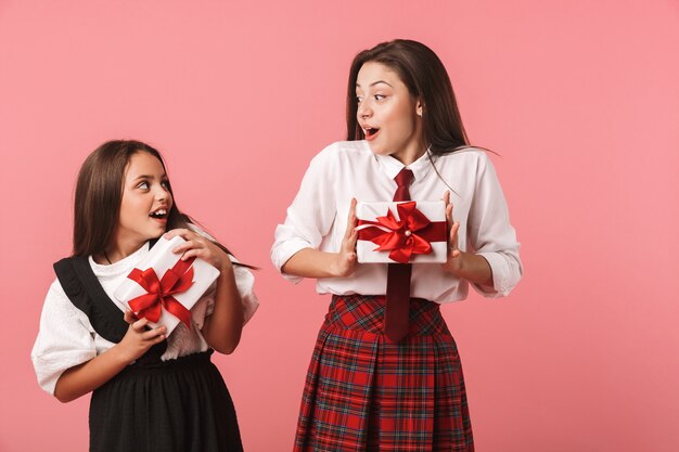 Portrait de filles joyeuses en uniforme scolaire tenant des boîtes présentes, tout en se tenant isolé sur mur rouge