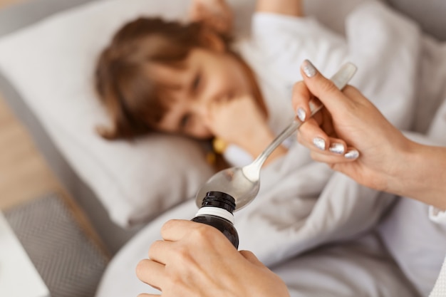 Portrait Flou D'une Petite Fille Avec Un Mal De Gorge Allongée Dans Son Lit, Une Mère Inconnue Sans Visage Donnant Du Sirop Contre La Toux, Une Famille Posant à L'intérieur à La Maison, Des Soins De Santé Pour Enfants Et Des Traitements.