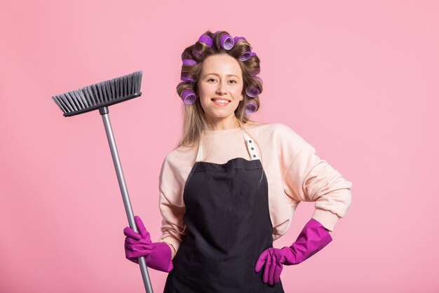 Portrait sur fond rose d'une femme souriante heureuse avec des rouleaux tordus dans les cheveux avec des gants de protection pour le nettoyage et un balai pour balayer le sol Femme de ménage travaillant après les heures de travail domestique