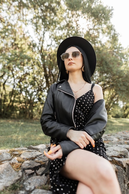 Portrait frais belle femme dans des lunettes de soleil à la mode dans un chapeau élégant dans des vêtements d'été noirs à la mode sur la nature par une journée ensoleillée. Fille élégante en tenue de mode est assise sur des pierres dans le parc. Vêtements de printemps pour femmes