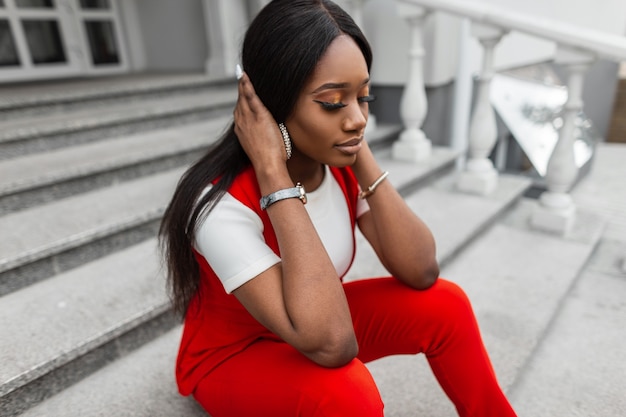 Portrait frais charmante jeune femme africaine modèle en costume à la mode rouge en ville. Belle fille noire avec des lèvres sexy dans des vêtements à la mode redresse la coiffure assise sur un escalier en pierre dans la rue.