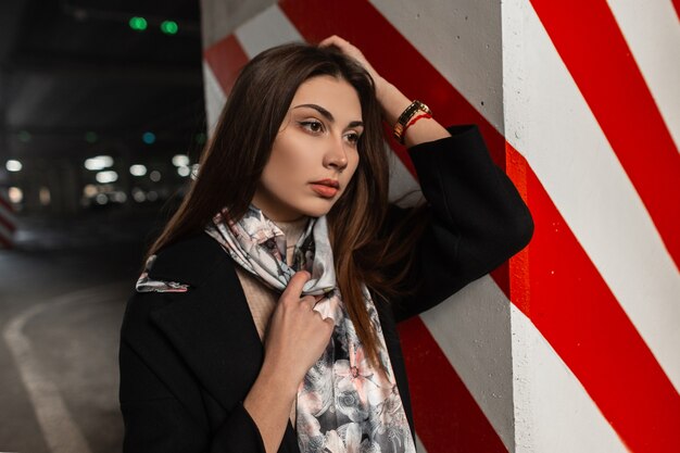 Portrait frais jolie jeune femme en manteau noir élégant en châle de soie à la mode près d'un pilier rouge-blanc moderne dans la rue près de la route. Beau modèle de femme élégante dans des vêtements à la mode dans la ville.