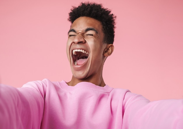 Portrait d'un garçon adolescent africain souriant portant un pull debout isolé sur rose, prenant un selfie, criant