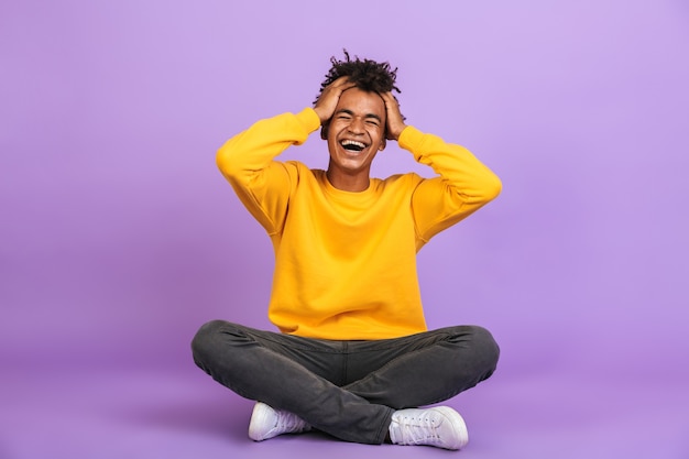 Portrait d'un garçon afro-américain excité riant et saisissant la tête alors qu'il était assis sur le sol avec les jambes croisées, isolé sur fond violet