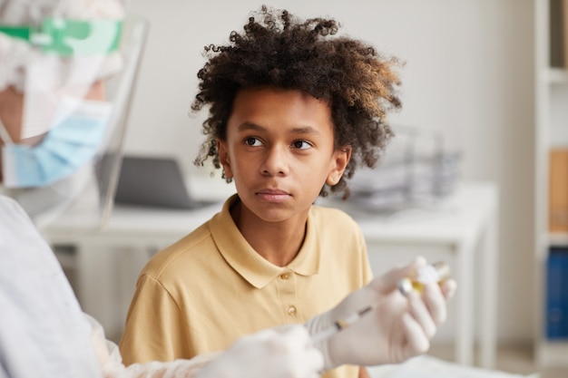 Portrait d'un garçon afro-américain regardant une infirmière dessinant un médicament dans une seringue pendant la vaccination contre le covid en clinique