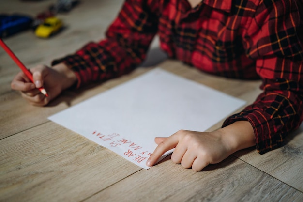 Portrait d'un garçon d'âge préscolaire en chemise à carreaux allongé sur le sol en écrivant une lettre de noël au père noël