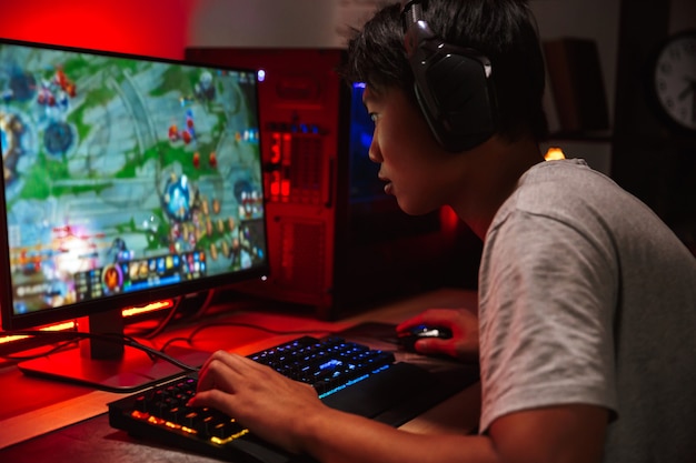 Portrait de garçon asiatique joueur jouant à des jeux vidéo sur ordinateur dans une pièce sombre, portant des écouteurs et à l'aide d'un clavier coloré rétroéclairé