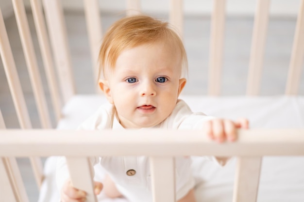 Portrait d'un garçon blond de 6 mois debout dans un berceau dans une chambre d'enfant lumineuse et souriant dans un body en coton le concept de produits pour enfants